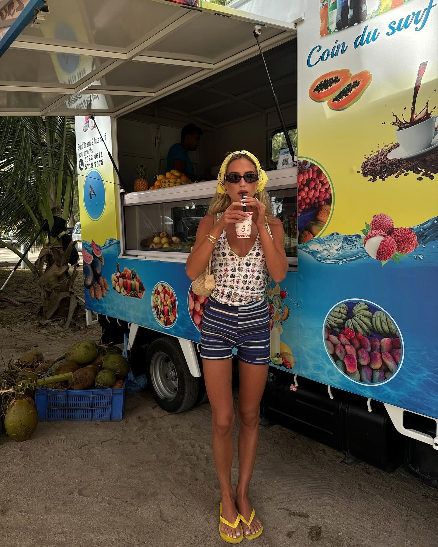 Mulher tomando um suco em frente a um food truck colorido na praia, rodeada de cocos no chão.