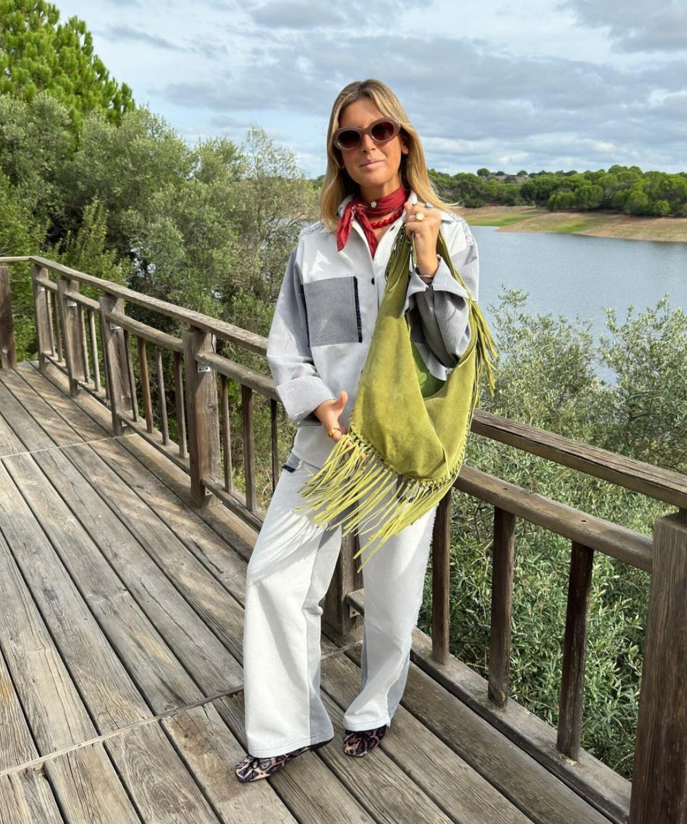 Mulher com óculos escuros e bolsa verde franjada, estilo acessórios boho, em uma varanda de madeira com vista para um lago.