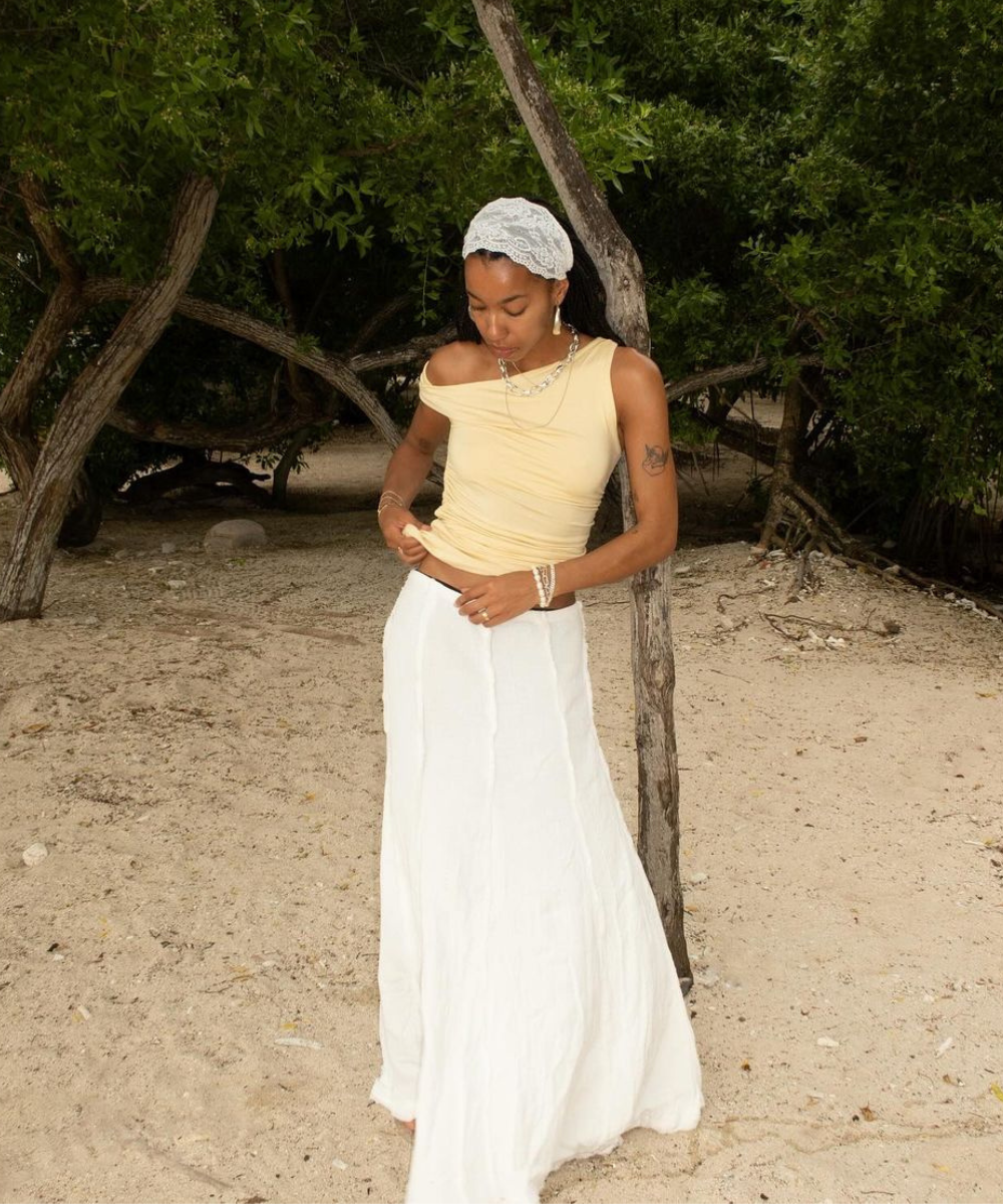 Mulher com saia longa e blusa amarela usa acessórios boho, incluindo um lenço e colares, em uma praia cercada por árvores.