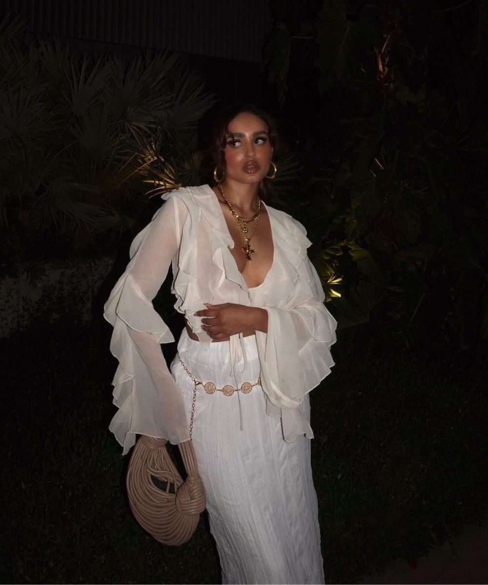 Mulher em vestido branco com babados, segurando bolsa trançada, usando acessórios boho em ambiente noturno.