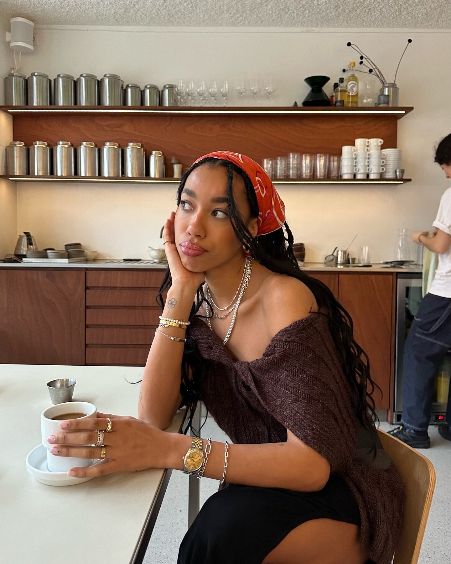 Pessoa com lenço vermelho na cabeça e várias joias, segura xícara de café em um café minimalista.