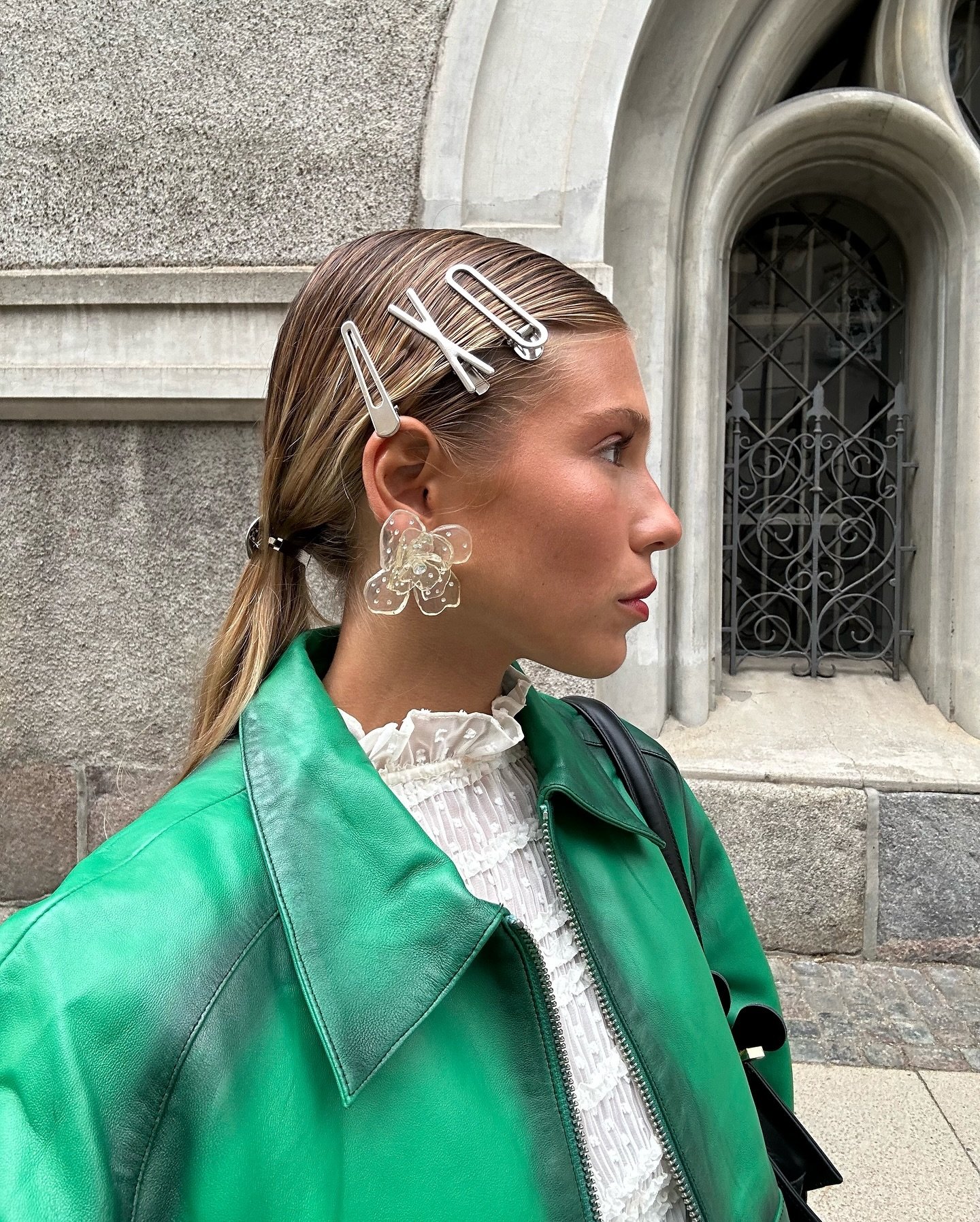 Mulher com prendedores metálicos no cabelo, usando brincos grandes e jaqueta verde. Acessórios de cabelo clássicos em destaque.