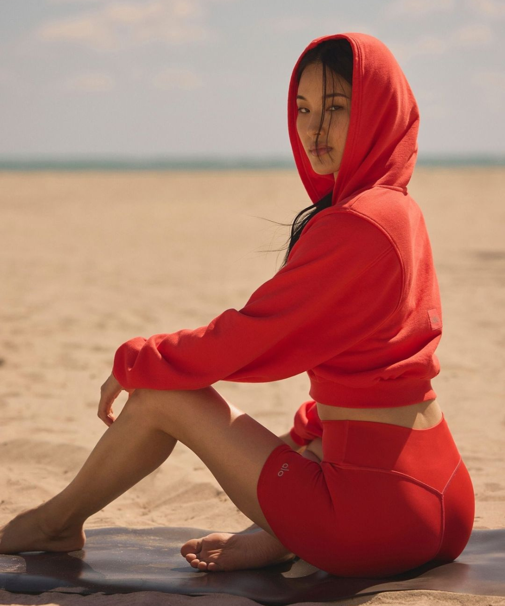 Pessoa sentada na praia usando roupas esportivas vermelhas ALO Yoga, olhando para a câmera, com o mar ao fundo.