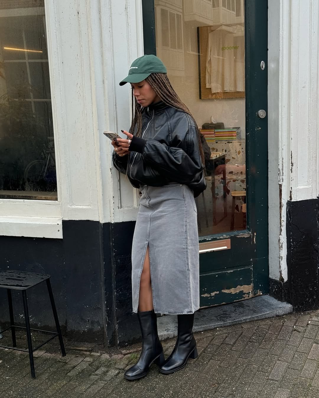 Mulher com jaqueta de couro, saia jeans e botas, segurando celular ao lado de uma loja.