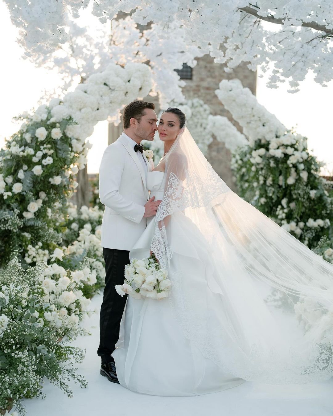 Casal elegante em cerimônia cercada por flores brancas, com a noiva usando um vestido luxuoso. Vestidos de noiva das celebridades.