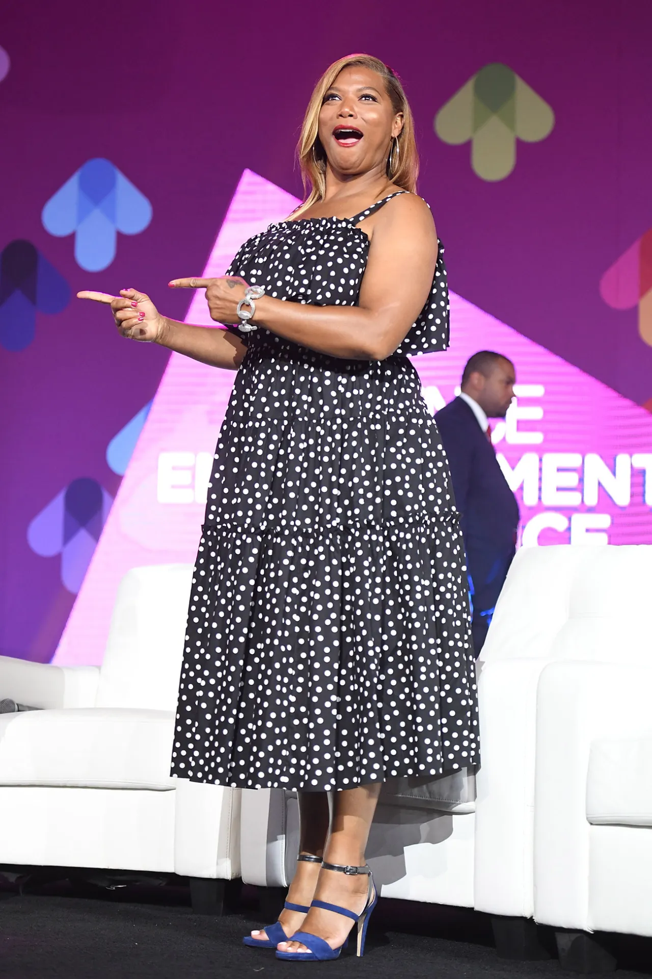 Pessoa com vestido de bolinhas e sandálias azuis, gesticula enquanto sorri em um evento em um palco iluminado.