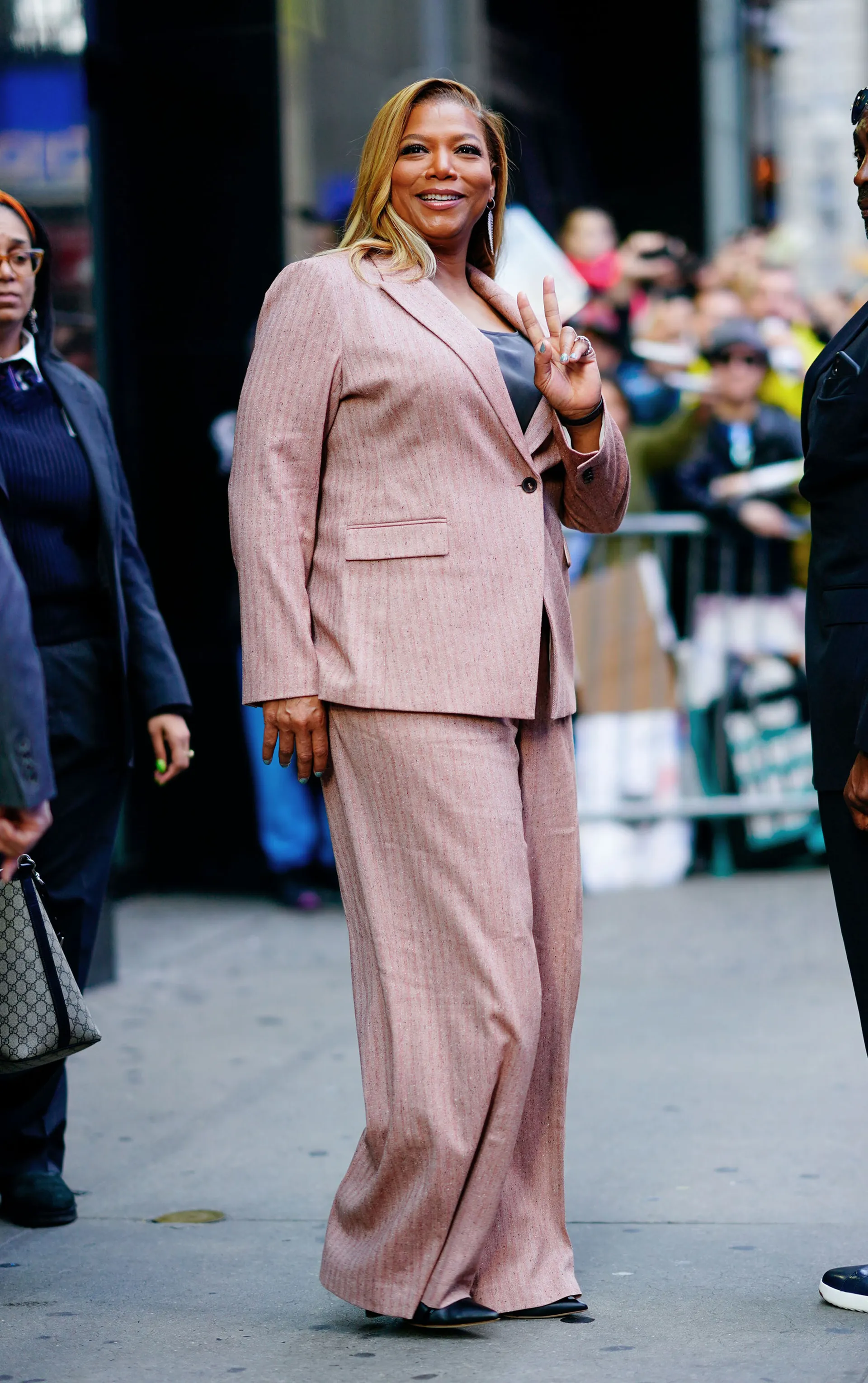 Queen Latifah em traje rosa claro faz sinal de paz, cercada por uma multidão.