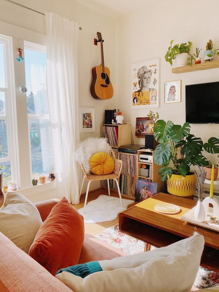 Sala de estar em apartamento pequeno com violão na parede, plantas, sofá laranja e poltrona com almofada amarela.