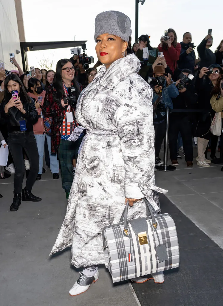 Queen Latifah com casaco estampado, chapéu cinza e bolsa xadrez em evento com fotógrafos.