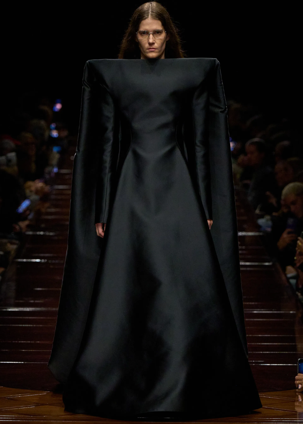 Modelo em passarela usando vestido preto volumoso, evocando estilo Romance Dark. Atmosfera sombria ao redor.