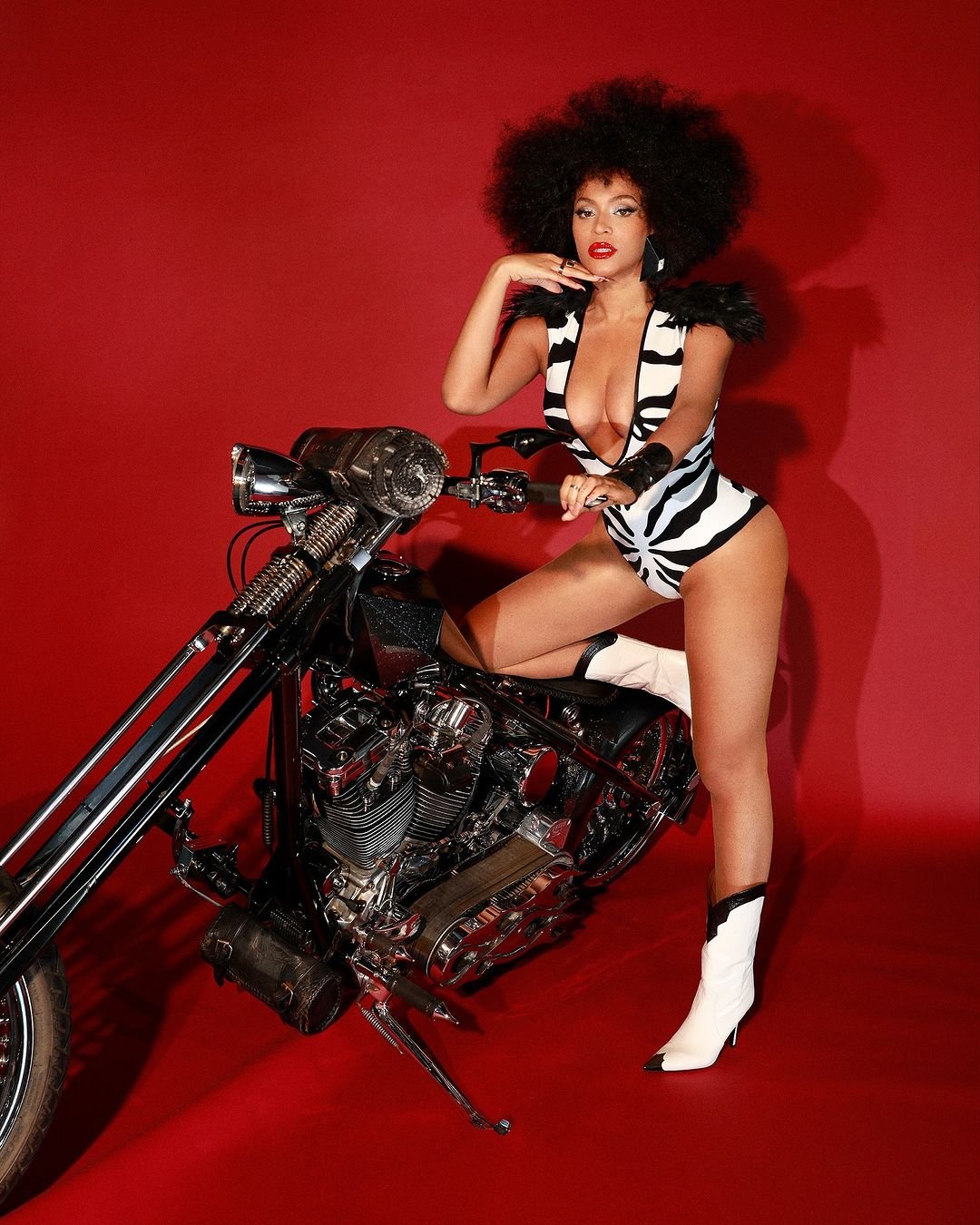 Mulher com cabelo afro em fantasia listrada, posa sobre moto preta contra fundo vermelho. Tema Halloween 2024.