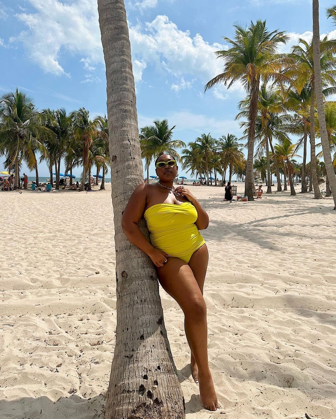 Pessoa em traje de banho e biquini para seios grandes amarelo encostada em coqueiro, em uma praia com palmeiras ao fundo.