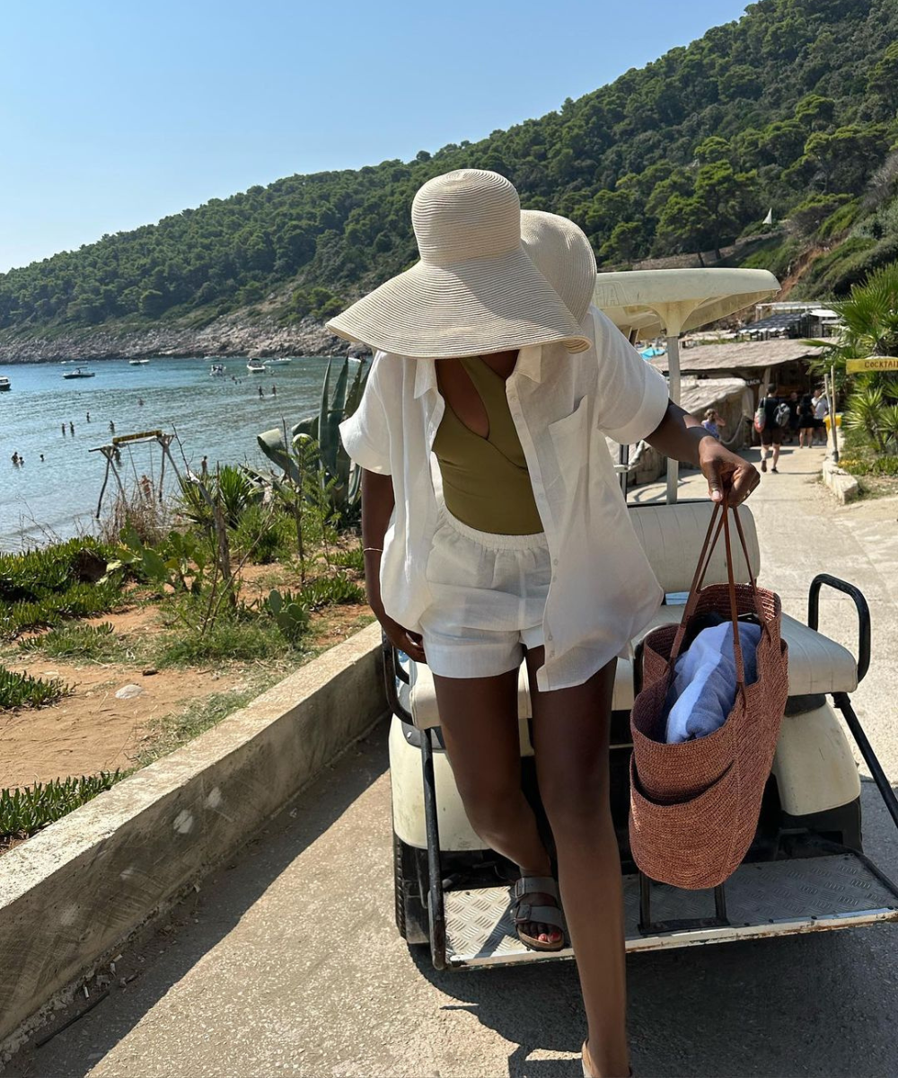 Mulher com chapéu grande segura bolsas de praia marrons perto do mar e vegetação.