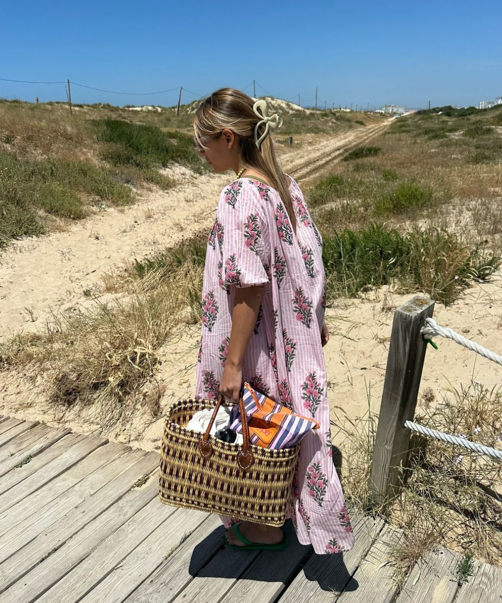 Mulher em vestido floral rosa caminha sobre passarela de madeira na praia, carregando bolsa de palha com toalha colorida.