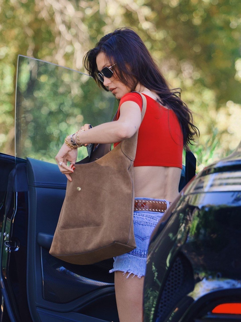 Mulher com camiseta vermelha e óculos escuros segura uma bolsa estilosa marrom ao lado de um carro.