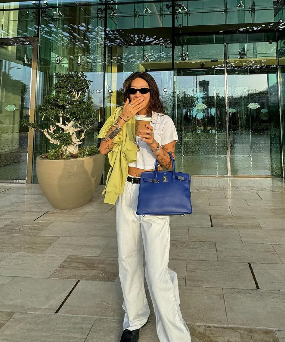 Mulher sorrindo com óculos de sol, camiseta branca e bolsa azul em ambiente moderno.