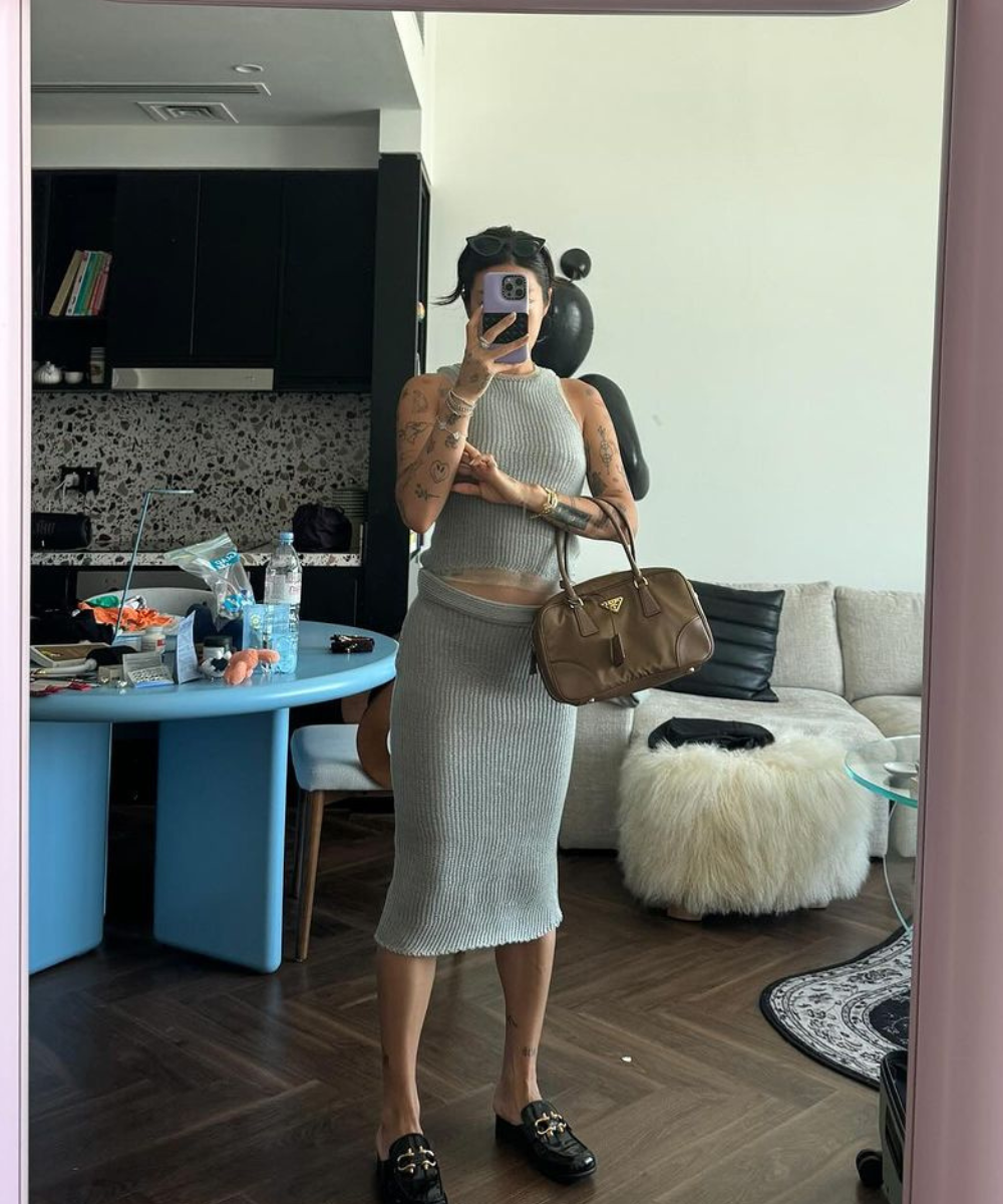 Pessoa com vestido cinza tira selfie no espelho, segurando bolsa marrom. Ambiente interno com sofá e mesa ao fundo.