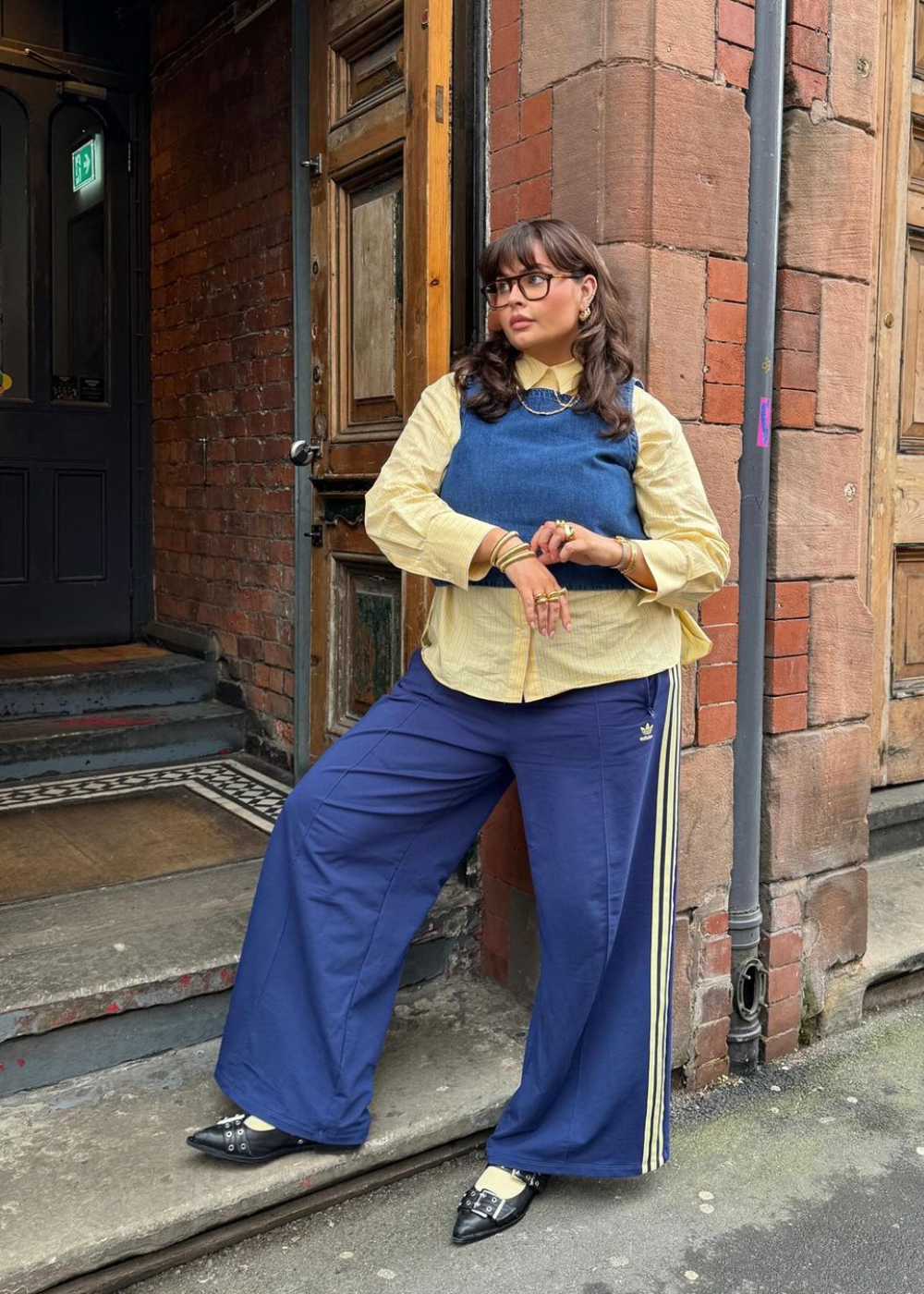 Pessoa usando calça com sapatilha preta, blusa amarela e colete azul-marinho, em frente a uma porta de madeira.