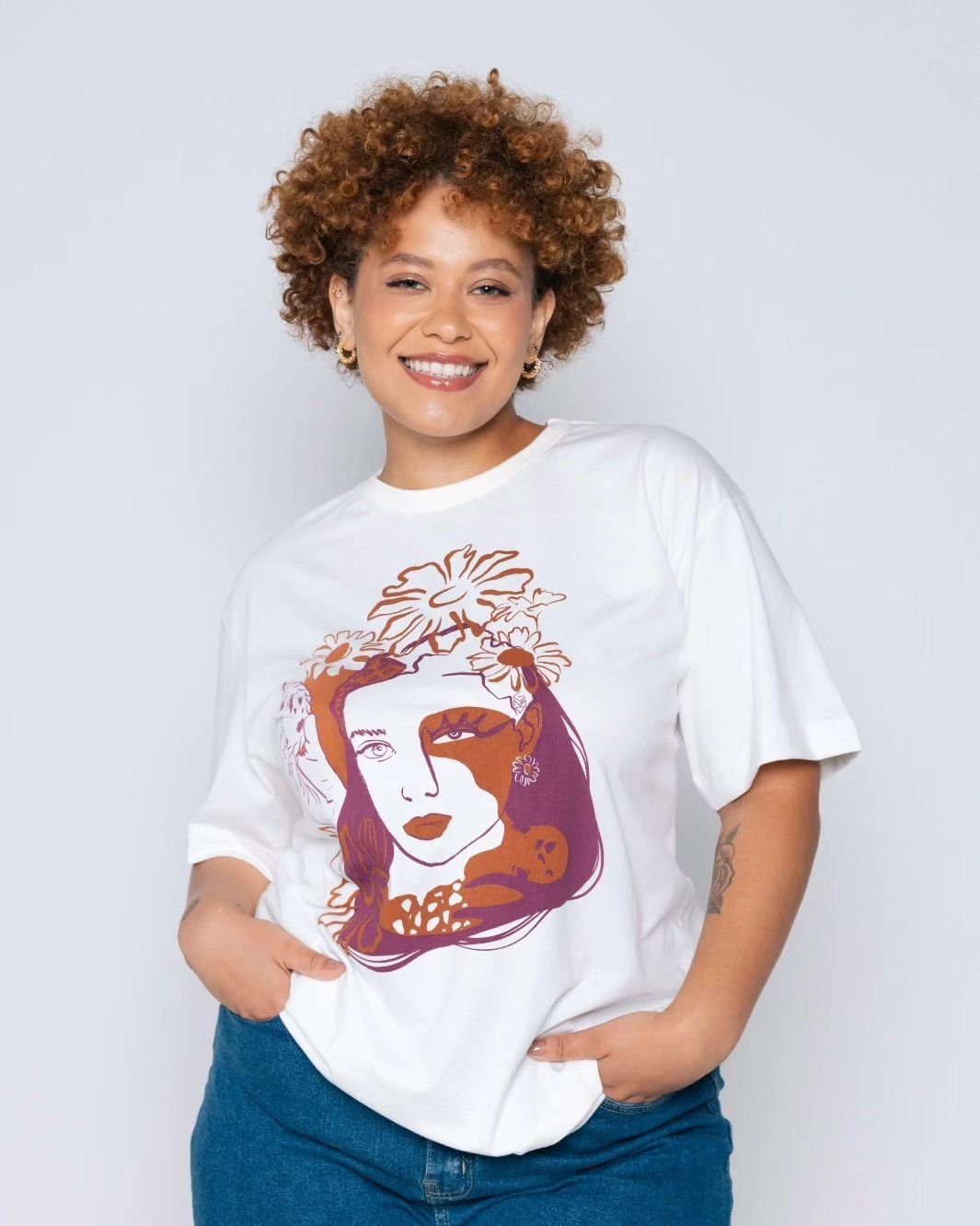 Pessoa sorridente com camiseta branca estampada com arte abstrata e floral. A camisa tem tons de marrom e rosa.