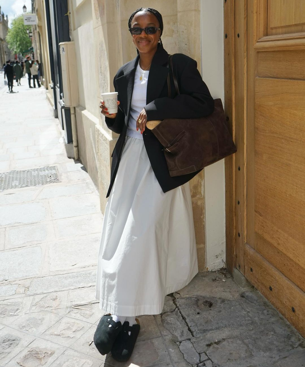 Pessoa com casaco preto e saia branca, segura café e uma bolsa de camurça. Está encostada em uma parede na rua.