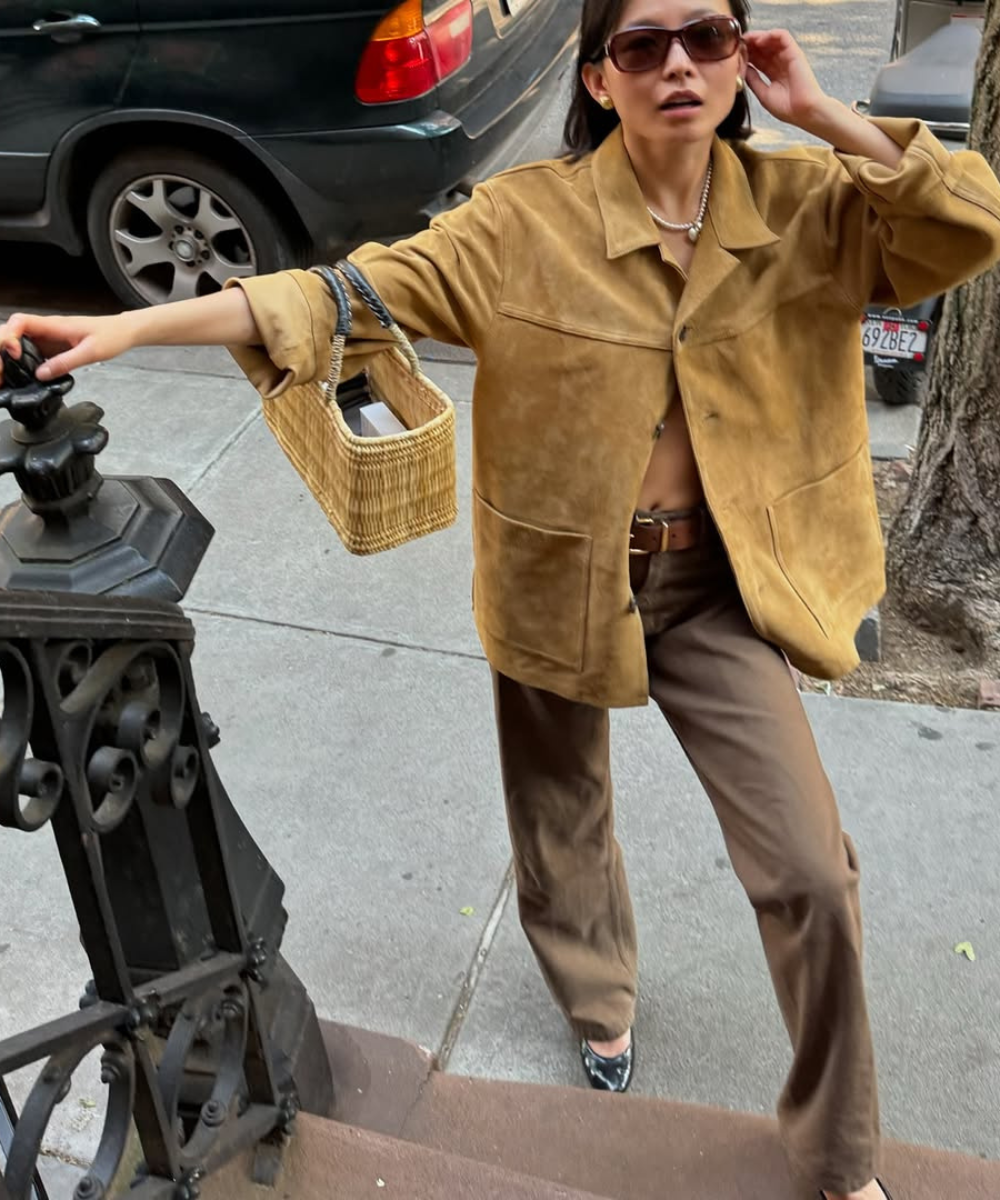 Pessoa em calçada usando jaqueta de camurça e calça marrom, carregando bolsa de palha e usando óculos escuros.