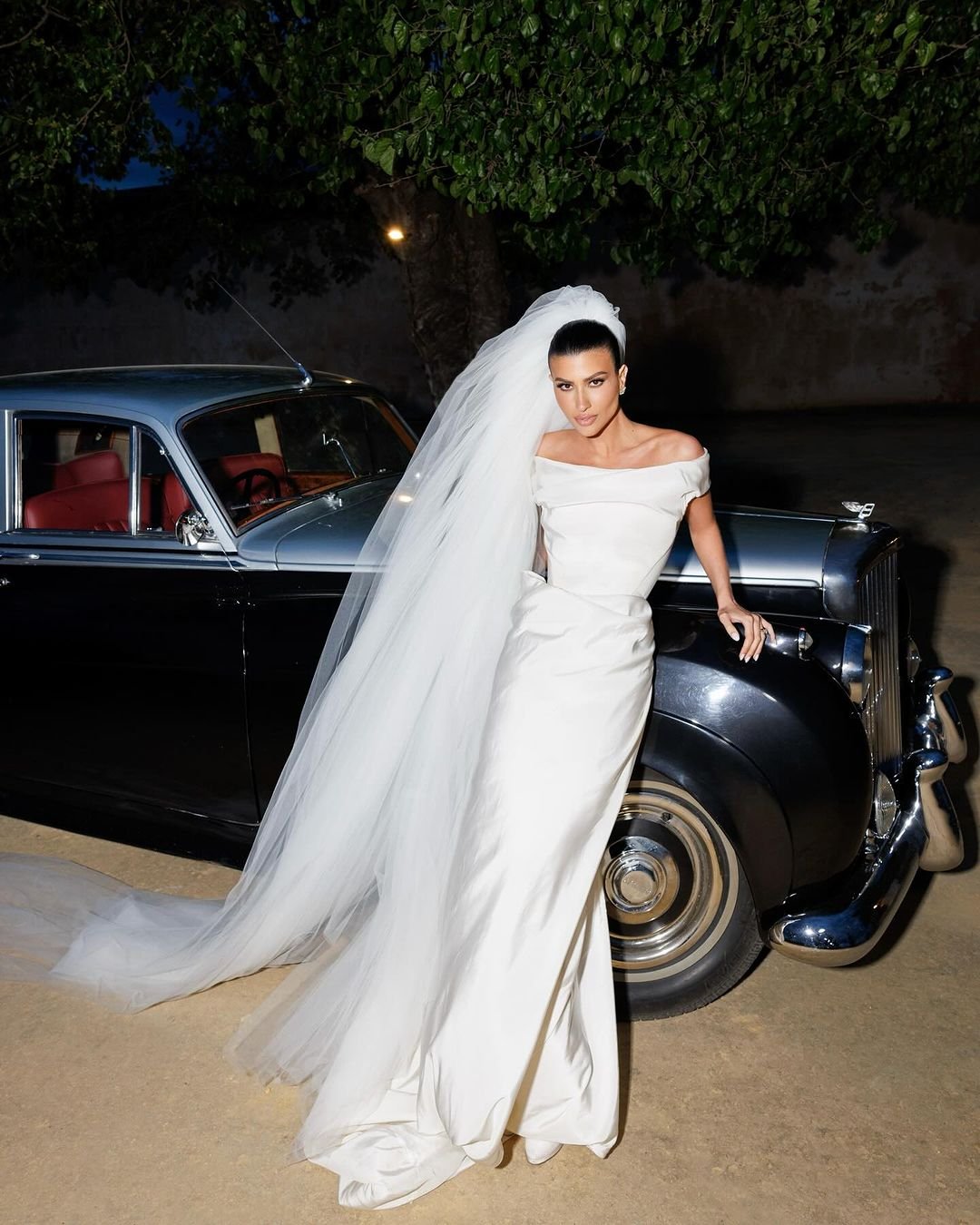 Noiva posando ao lado de carro clássico, com vestido elegante e véu longo, representando o glamour dos vestidos de noiva das celebridades.