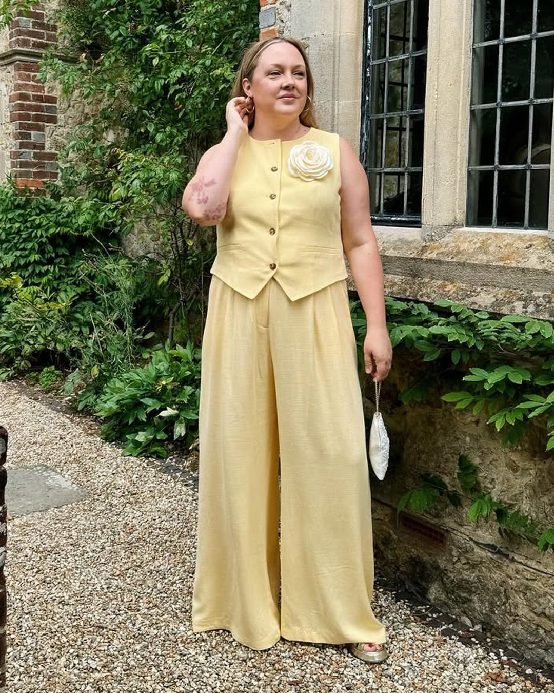 Pessoa em um conjunto amarelo claro elegante, com uma pequena bolsa de mão moderna e um broche de flor.