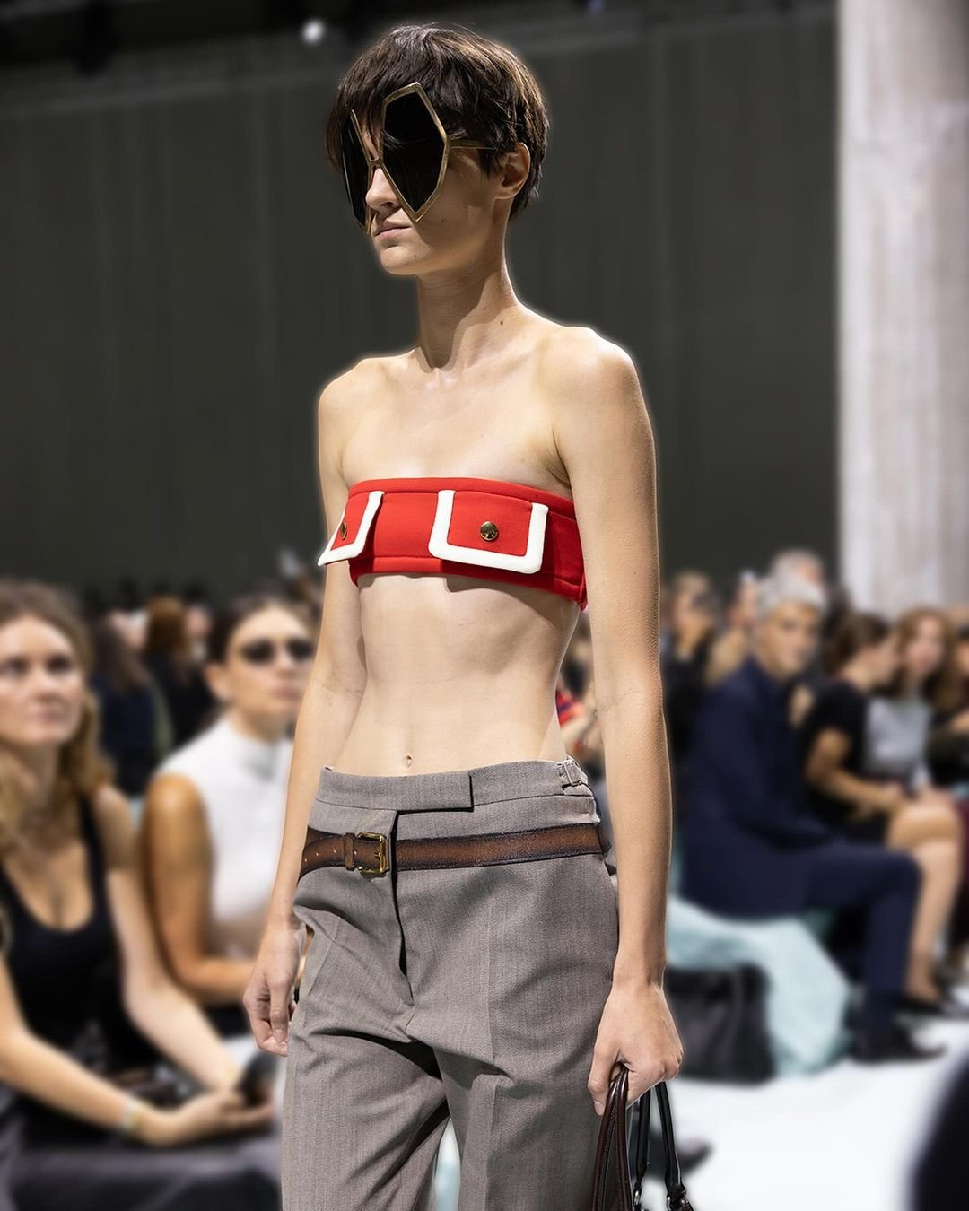 Modelo usando calça cinza com cinto em destaque, combinando com top vermelho em desfile.