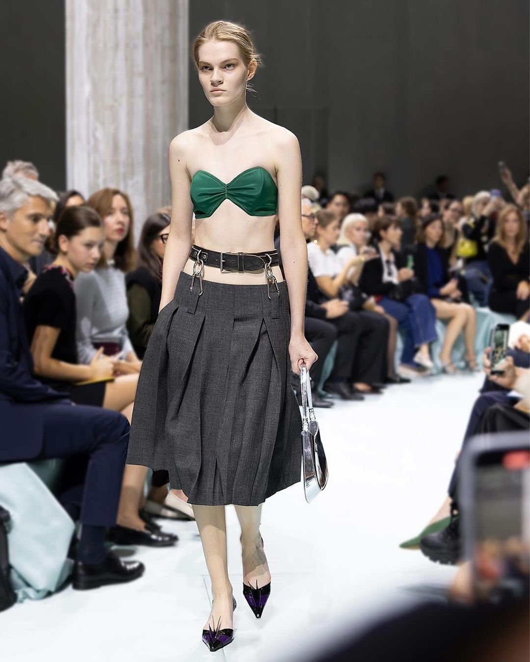 Modelo exibindo saia cinza e top verde em desfile, com cintos tendência e bolsa prateada.