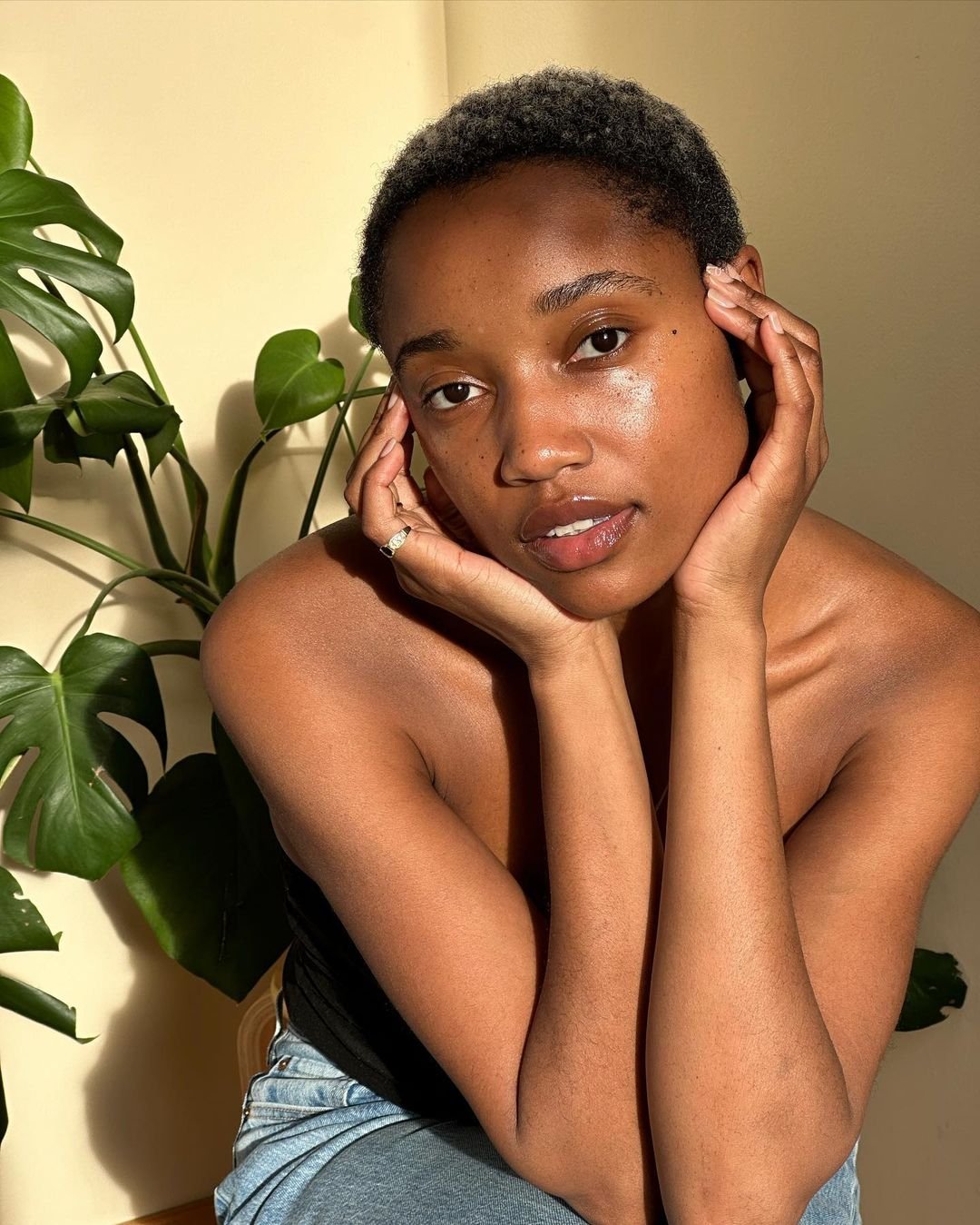 Jovem mulher de cabelo curto em ambiente iluminado, com plantas ao fundo, transmite serenidade.