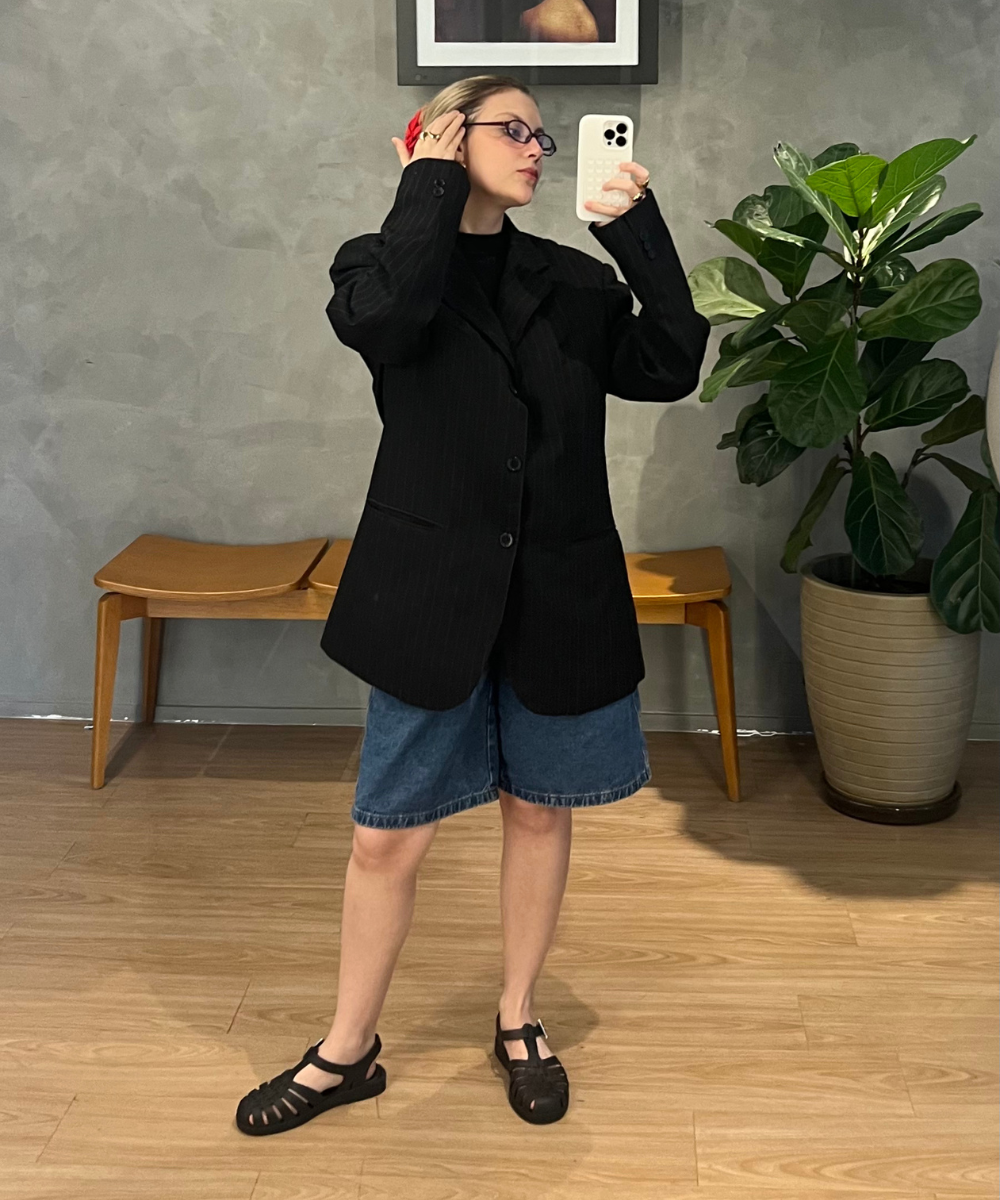 Pessoa com blazer preto e bermuda jeans tira selfie, usando sandálias estilo Melissa. Ambiente moderno com planta ao fundo.