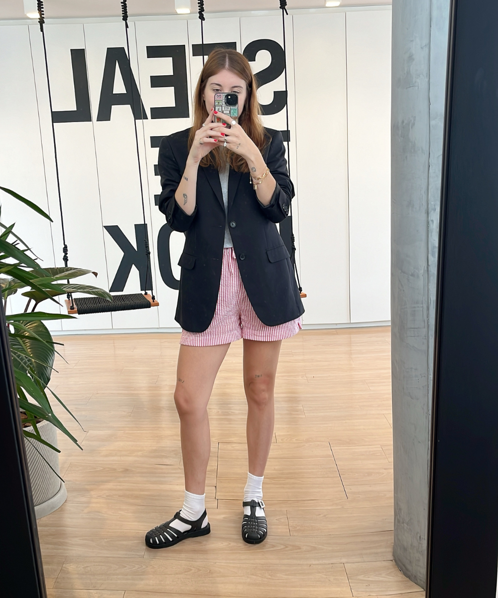 Mulher com blazer preto, shorts listrados e sandálias Melissa, tirando selfie em ambiente moderno. Mostra como usar looks com melissa no escritório.