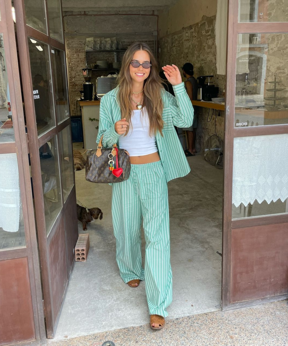 Mulher sorridente saindo de uma cafeteria, usando um conjunto listrado verde e sandálias fisherman.