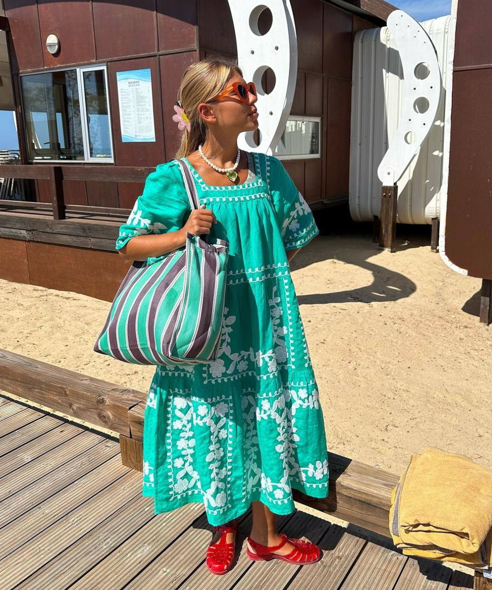 Vestido verde com bordado floral branco, bolsa listrada e sandália fisherman vermelha na praia.