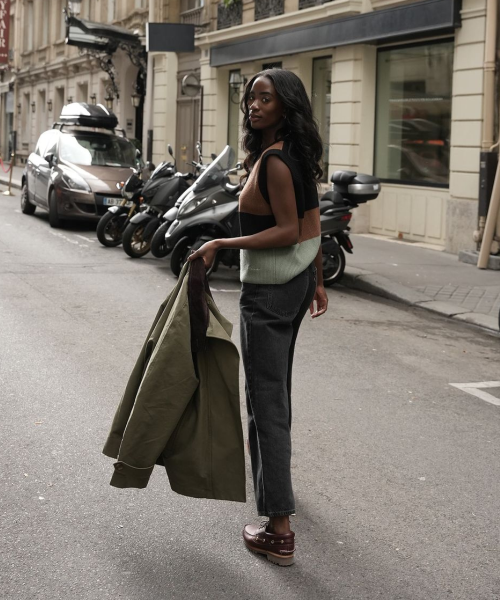 Mulher de colete multicolorido e calça jeans na rua, segurando casaco. Calçados dockside marrons.