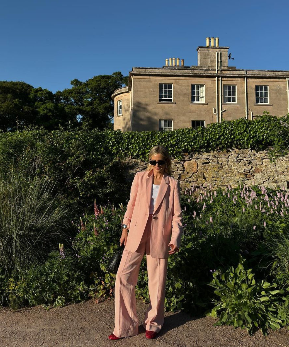 Mulher em terno rosa claro elegante, em frente a uma casa clássica, cercada por plantas.