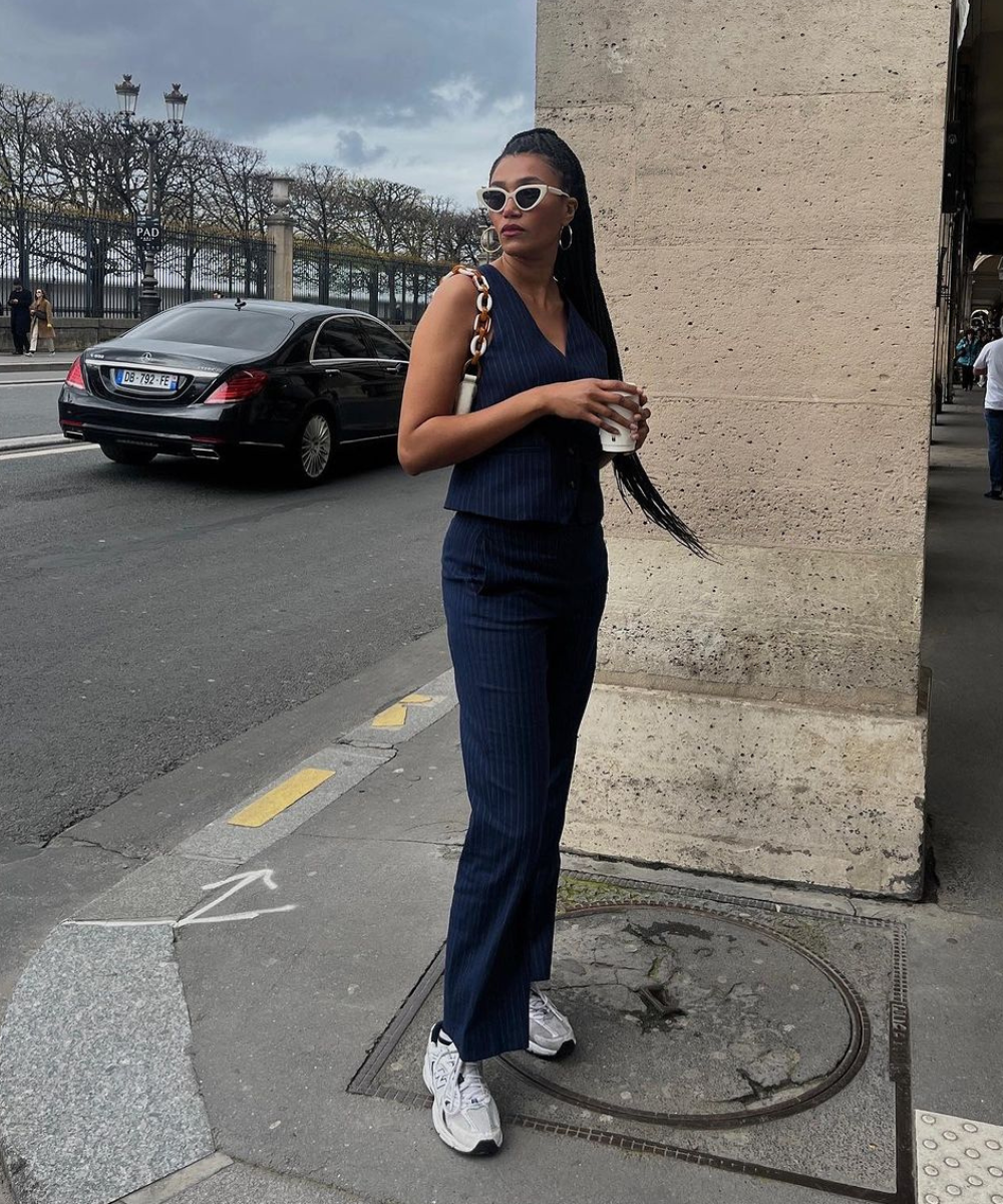 Pessoa elegante em look azul escuro, com óculos de sol e tênis, em rua urbana. Cores tendência chiques realçam o estilo.