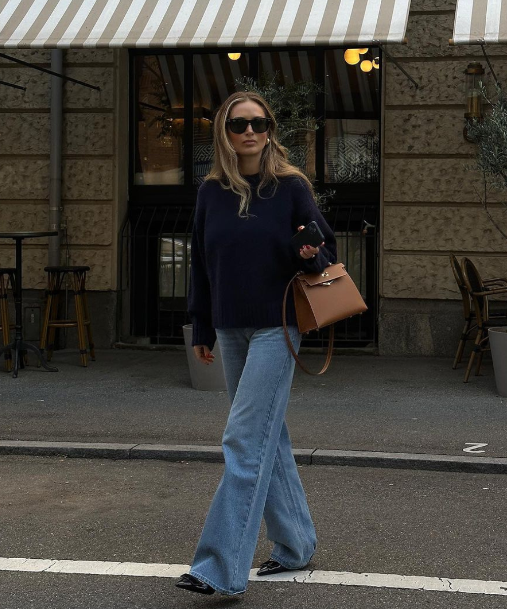 Mulher vestindo suéter azul e jeans flare, com bolsa marrom, caminhando em frente a um café. Elegância casual em tons chiques.