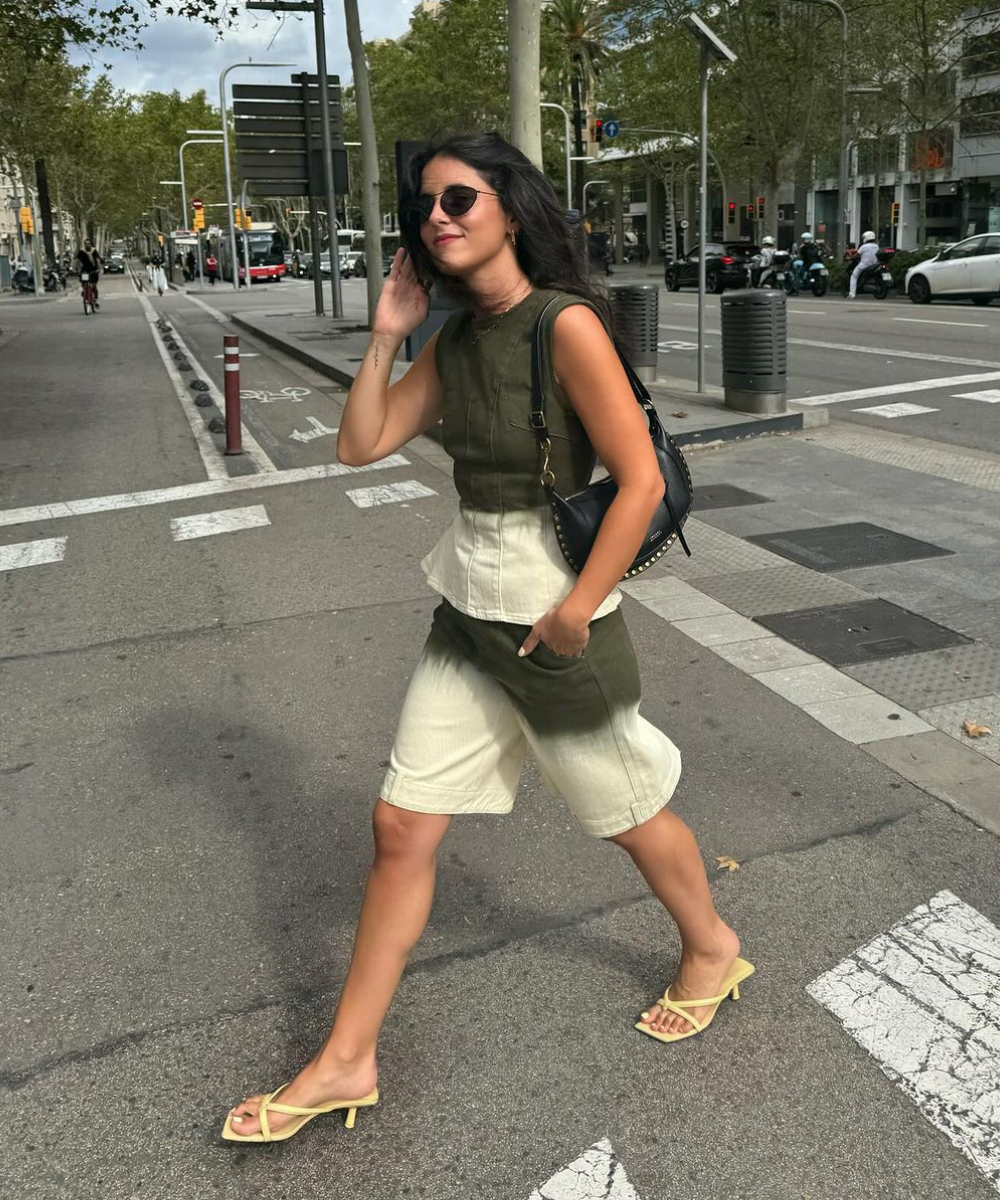 Mulher atravessa a rua com conjunto verde e bege, sandálias amarelas e bolsa preta. Estilo casual e moderno.