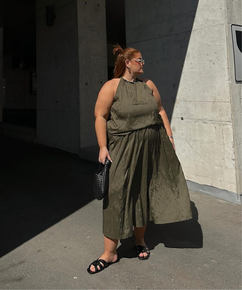 Mulher com vestido verde oliva em cenário urbano, segurando bolsa preta, envolve tendência em cores chiques.