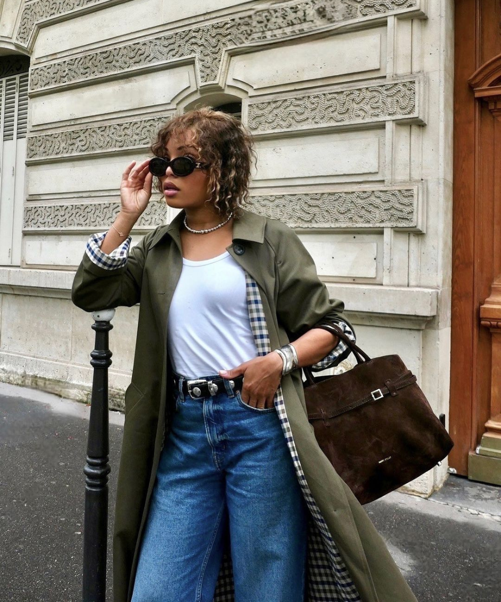 Mulher com casaco verde e jeans, segurando óculos escuros. Bolsas e acessórios destacam cores tendência chiques.