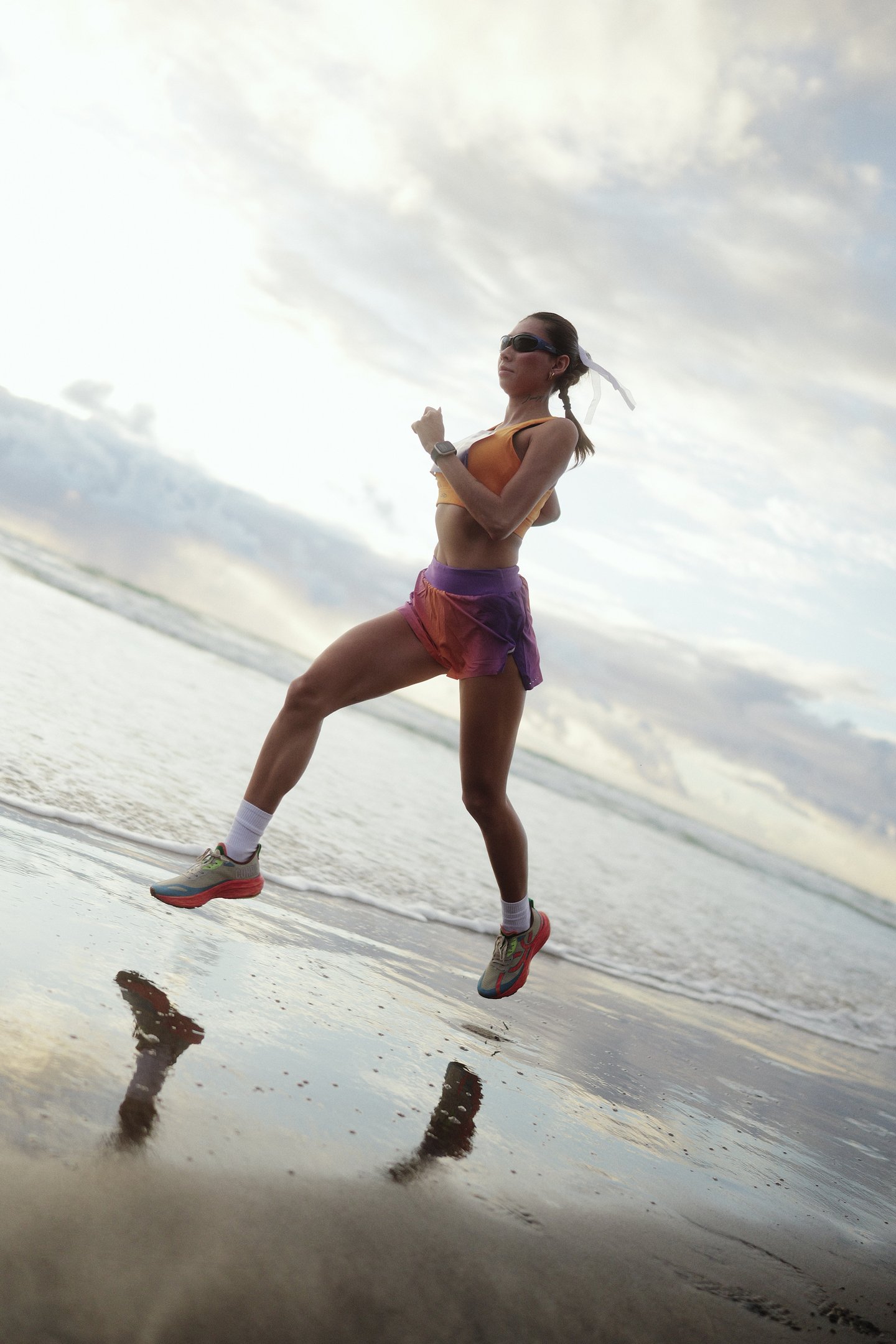 Pessoa correndo na praia ao amanhecer, usando óculos escuros e roupa esportiva colorida. Tênis de oncinha.