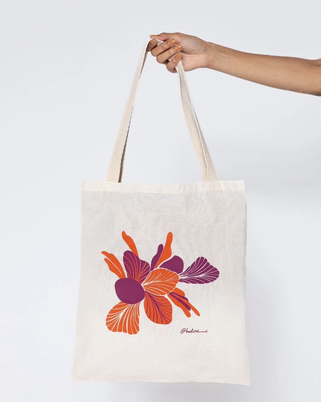 Sacola de tecido bege com estampa floral em laranja e roxo segurada por uma mão. Ideal para compras ou uso casual.