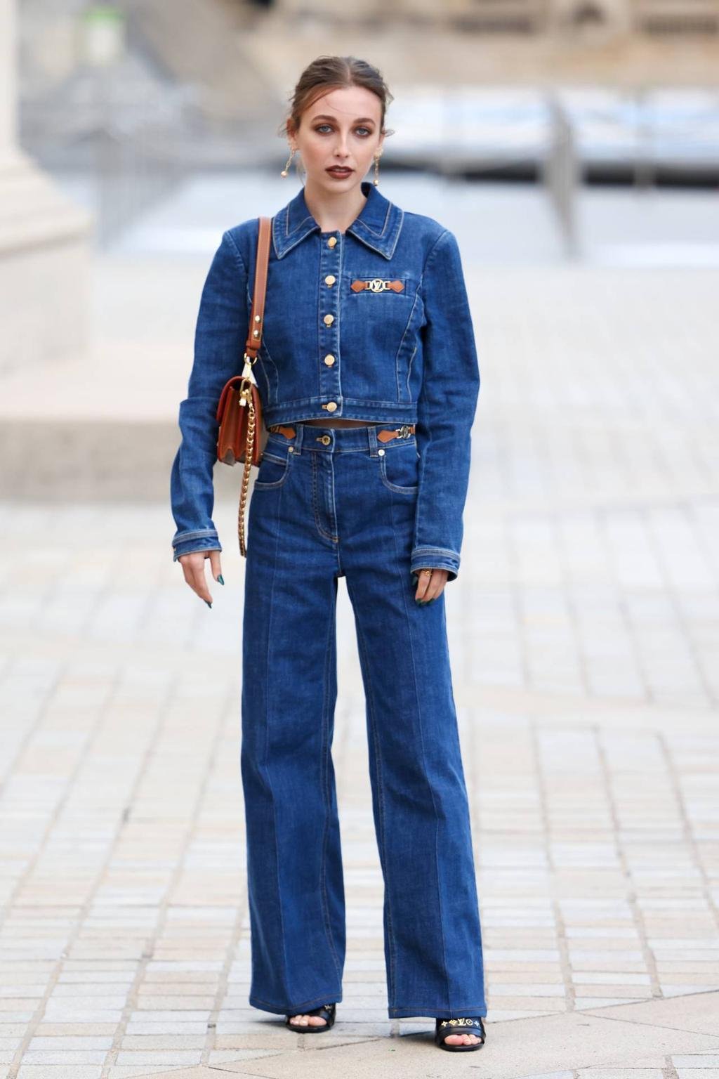 Looks da Emma Chamberlain em conjunto jeans estiloso e bolsa marrom em local urbano, exemplificando looks modernos e despojados.