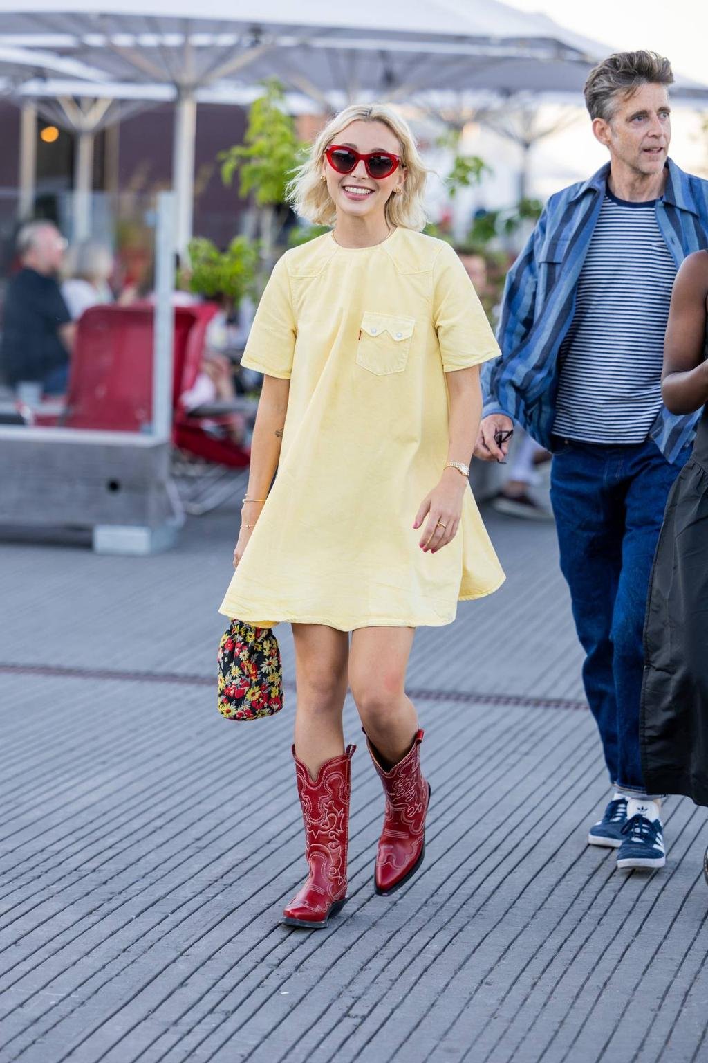 Looks da Emma Chamberlain usando vestido amarelo claro, botas vermelhas e óculos escuros em formato de coração, carregando uma bolsa florida.