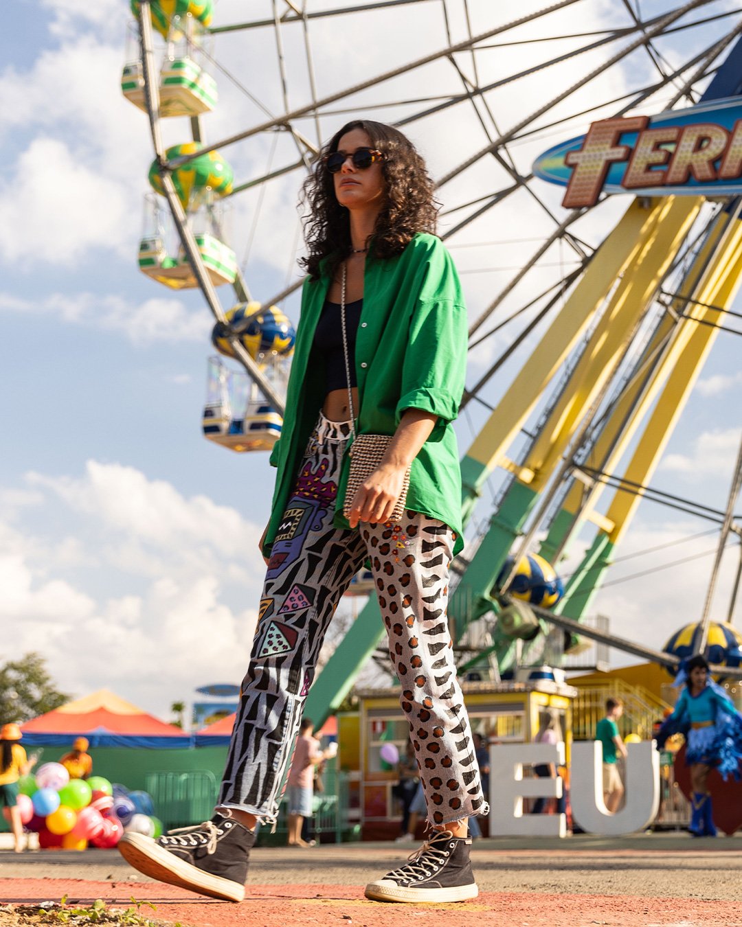 Pessoa de óculos escuros e cabelo cacheado veste camisa verde e calça estampada em parque de diversões com roda-gigante.