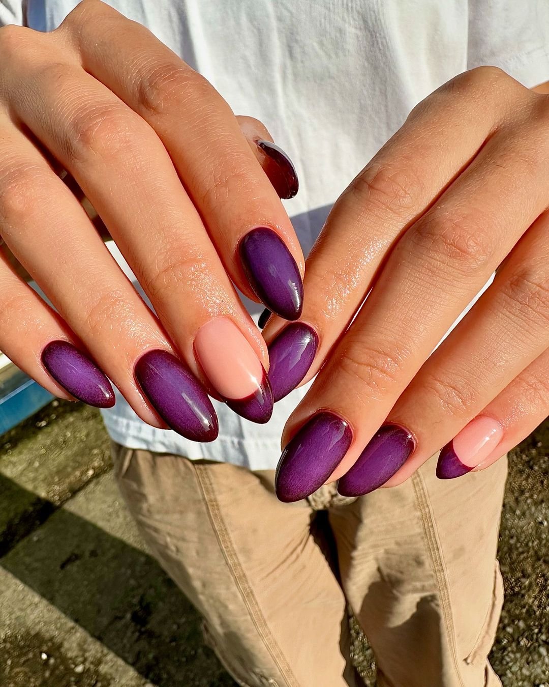 Mãos com esmalte roxo e nude, unhas em formato amendoado decoradas com estilo moderno.