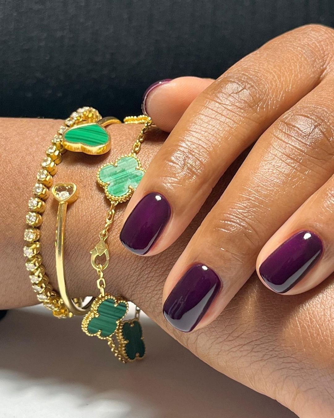 Unhas roxas bem cuidadas adornadas com pulseiras douradas e verdes elegantes.