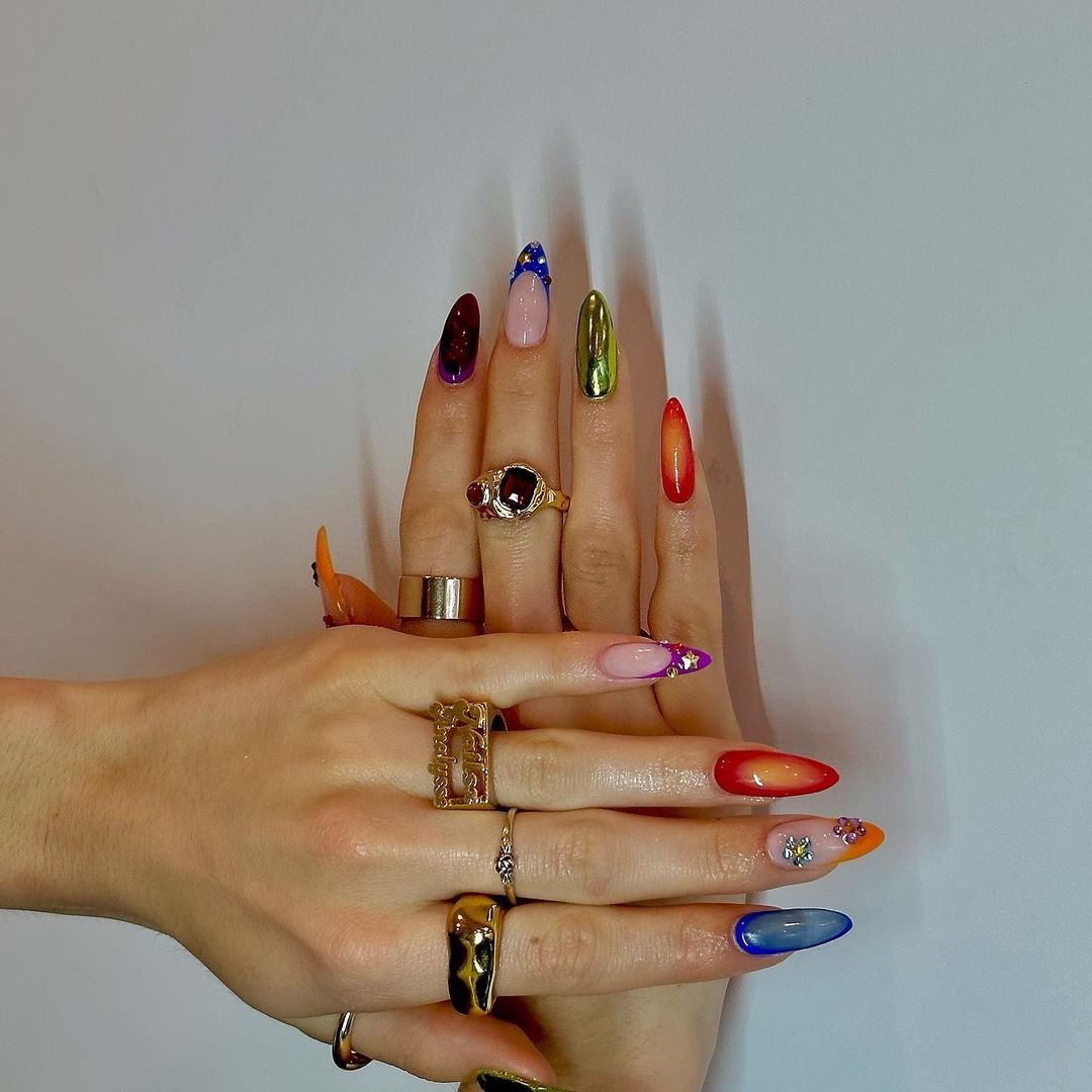 Unhas com designs coloridos e artísticos, alinhadas às tendências de manicure 2025. Mãos exibem vários anéis.