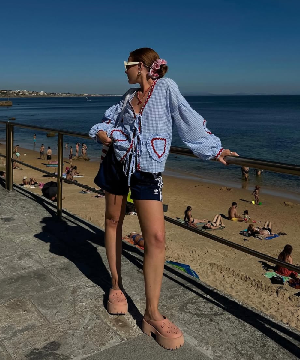 Pessoa usando casaco xadrez azul estilo vichy com corações vermelhos, em frente à praia ensolarada.